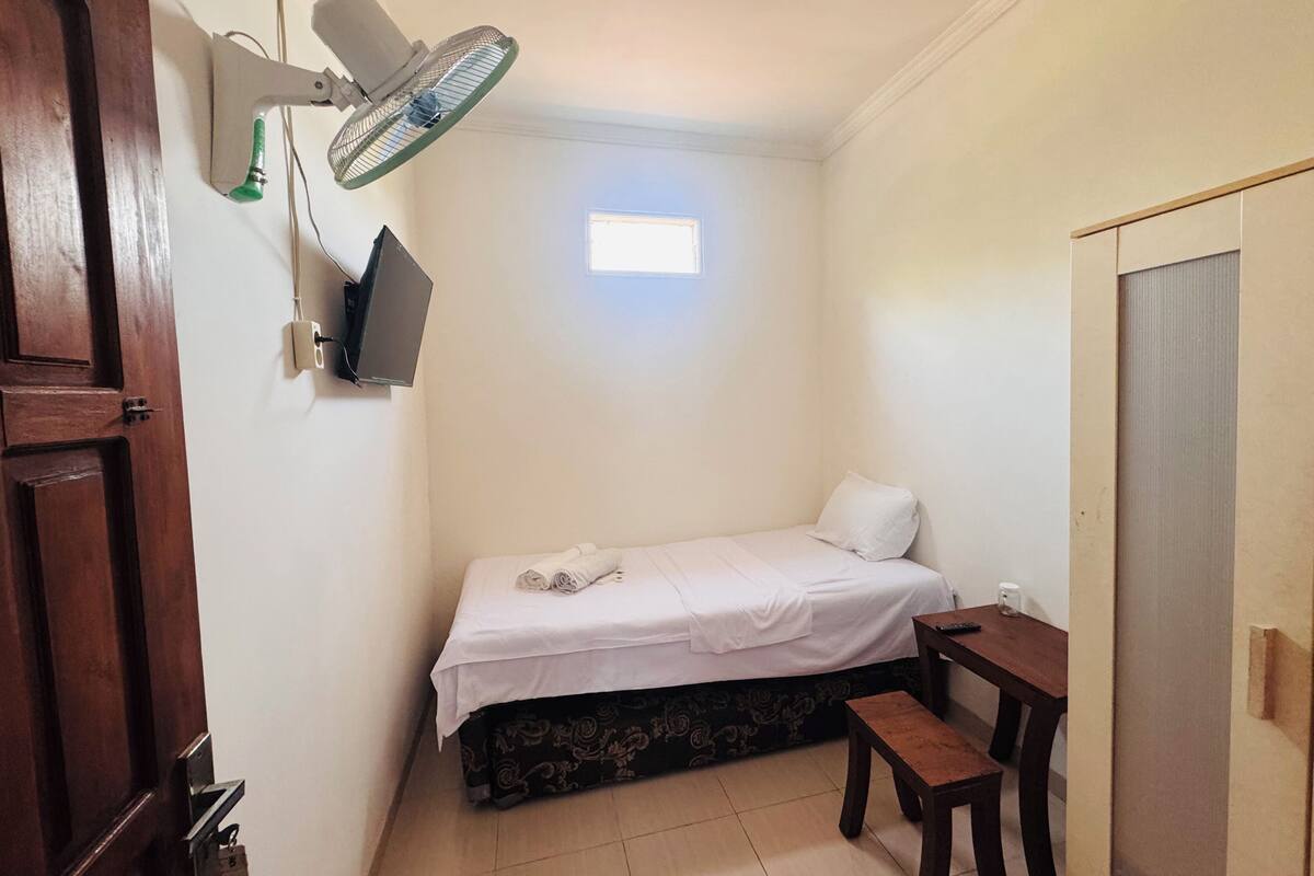 Un dormitorio pequeño y ordenado con cama, escritorio, silla, televisor montado en la pared y ventilador de techo.