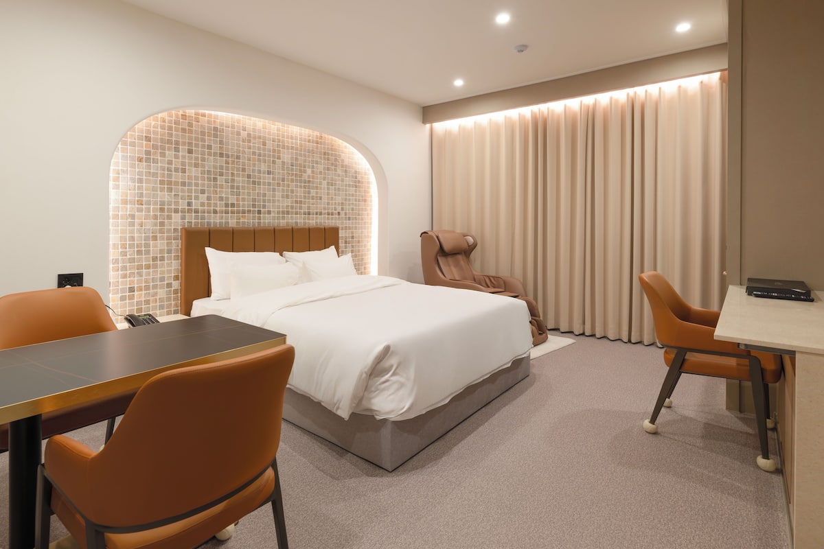 A modern hotel room with a neatly made bed, a massage chair, a desk, and a chair.