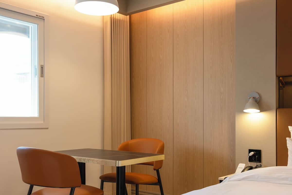 A hotel room with a bed, a table, two chairs, a desk, a lamp, and a window.