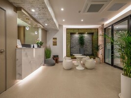 Un área de recepción moderna con una barra de mármol, mostrador de recepción, planta en la pared y zona de estar con mesas y sillas blancas.
