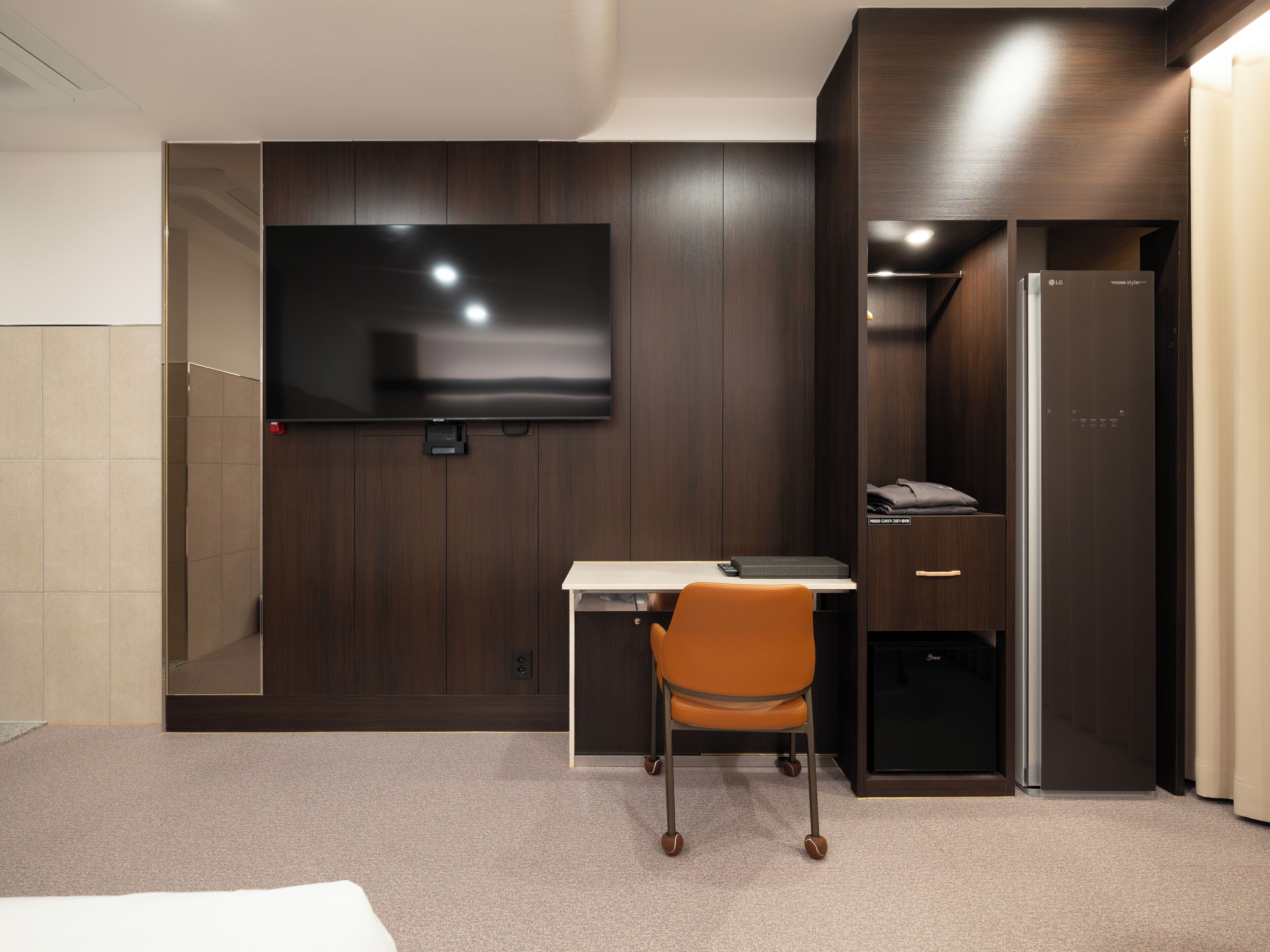 A modern hotel room with a flat-screen TV mounted on the wall, a desk with a chair, a wardrobe, and a minibar.