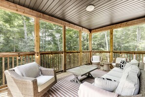 A covered outdoor seating area with wicker furniture, a glass table, and a view of a forest.