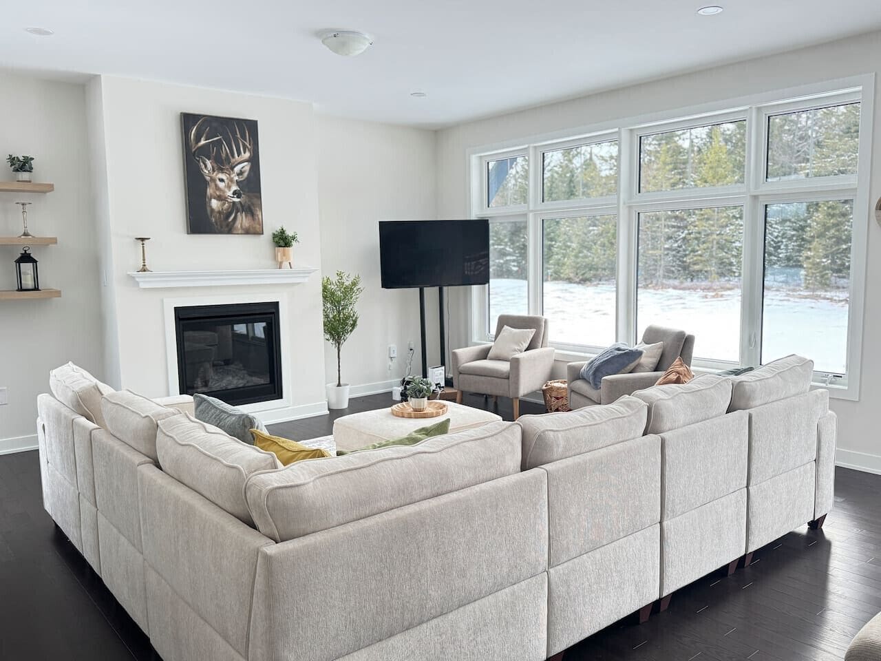 A modern living room with a sectional sofa, a fireplace, a flat-screen TV, and large windows offering a view of the outdoors.