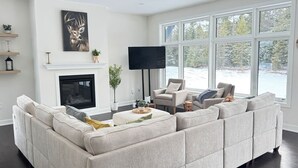 Una sala de estar moderna con un sofá modular, una chimenea, un televisor de pantalla plana y ventanas grandes que ofrecen vistas al exterior.