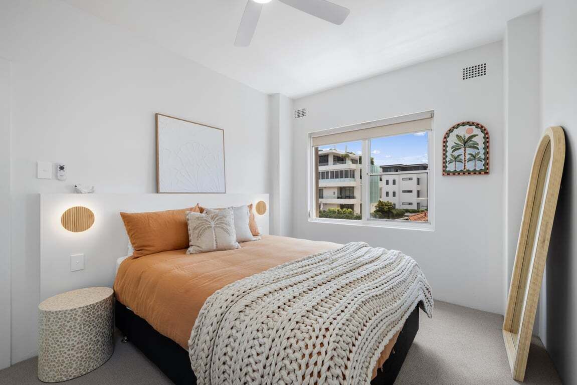 Une chambre à coucher avec un lit, une table de chevet ronde, une fenêtre donnant sur des immeubles et un tableau accroché au mur.