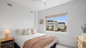 Un dormitorio con una cama, una mesita de noche con lámpara, una ventana con vistas a edificios y un ventilador de techo.