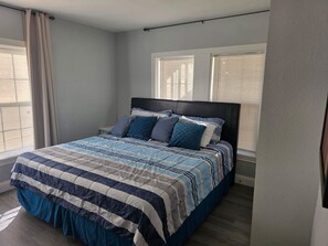 Ein Schlafzimmer mit einem Bett, zwei Fenstern mit Jalousien, einem Vorhang und einem Kopfteil.