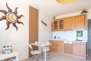 A modern kitchen with wooden cabinets, a white countertop, and a dining area with a white table and chairs. - L'appartement Cala Nova (BORMES LES MIMOSAS)