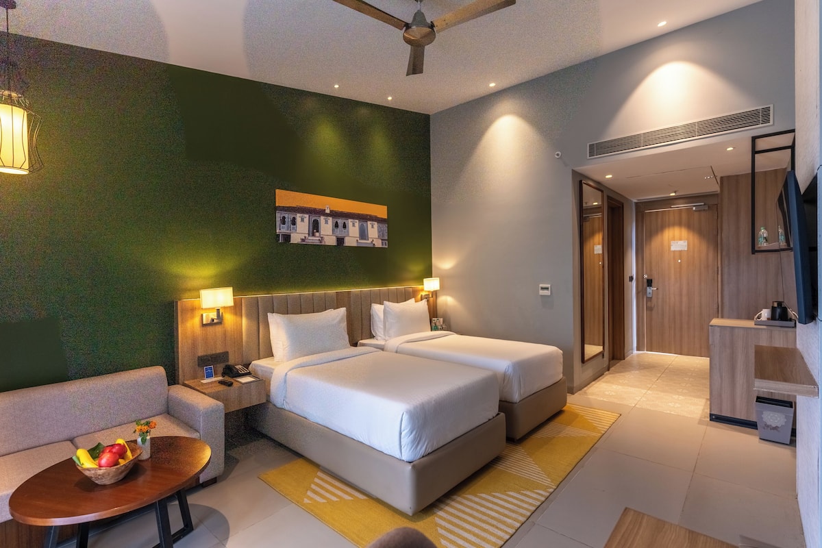 A hotel room with two beds, a sofa, a small table with fruit, and a wall-mounted TV.