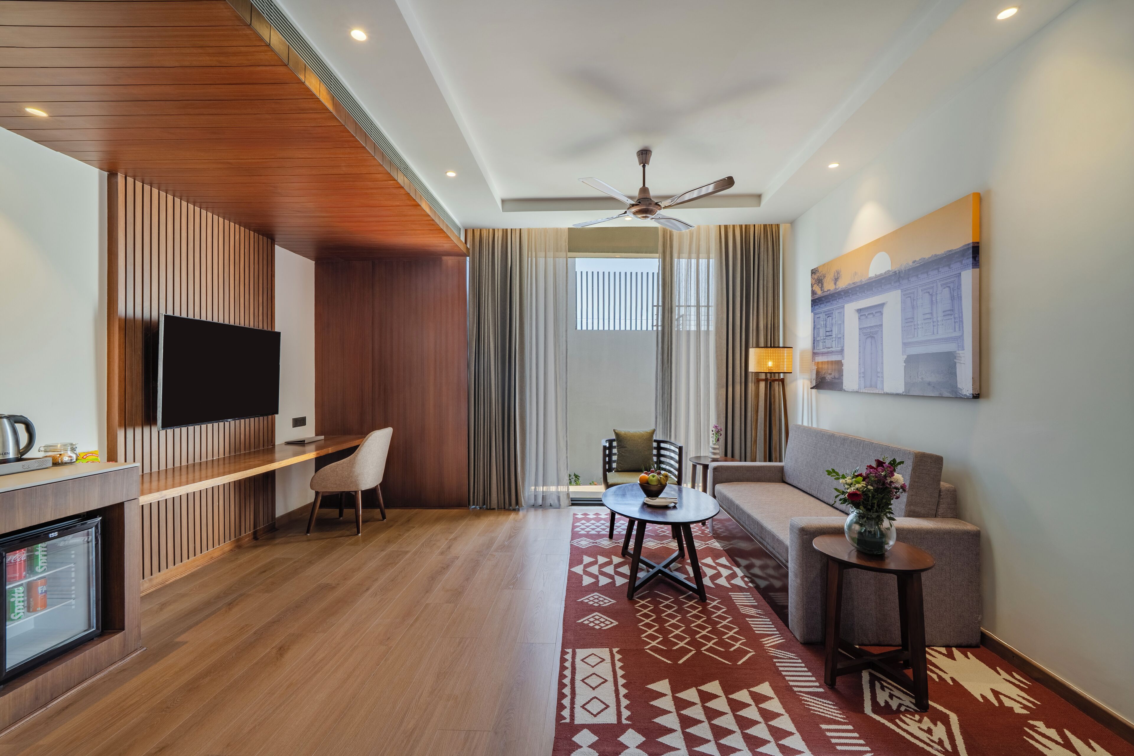 A hotel room with a wooden ceiling, a flat-screen TV, a small dining table, a sofa, and a patterned rug.