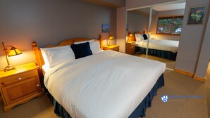 Ein ordentlich gemachtes Bett mit weißer Bettwäsche und blauer Bettdecke, zwei Nachttische mit Lampen, ein Spiegel an der Wand und eine gerahmte Karte.