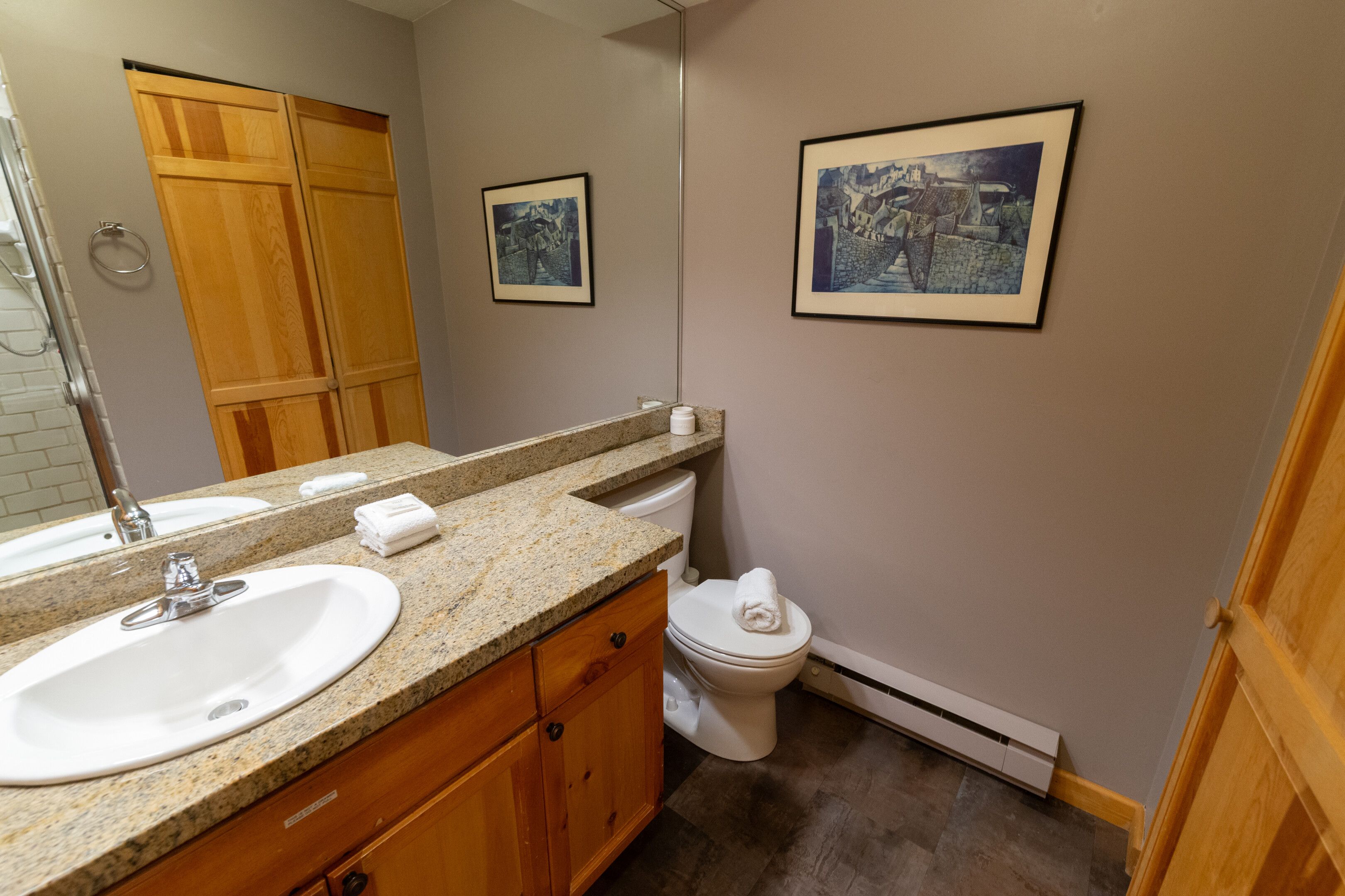 A bathroom with a granite countertop, white sink, wooden cabinets, a toilet, and framed pictures on the wall.