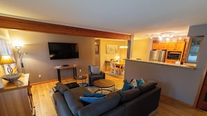 Ein Wohnzimmer mit Sofa, Couchtisch und Fernseher. Es gibt einen Essbereich mit Tisch und Stühlen. Die Küche ist sichtbar, mit Schränken und Geräten.