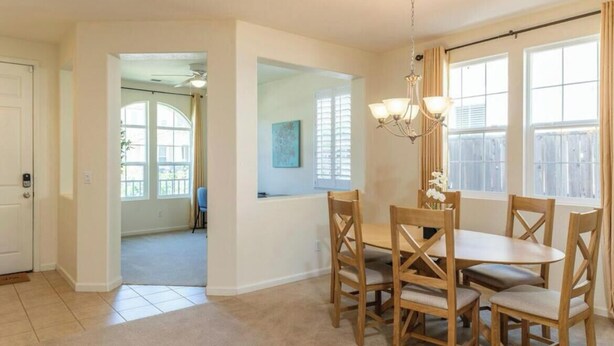 A dining area with a round table and wooden chairs, a chandelier, and large windows with curtains.