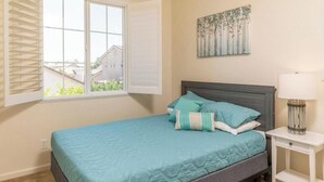 A bedroom with a bed, a window with blinds, a bedside lamp, and a wall-mounted artwork.