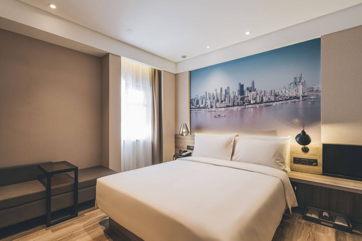 Ein modernes Hotelzimmer mit einem großen Bett, einer Stadtlandschaftsmalerei, einem Nachttisch mit Lampe und Holzboden.