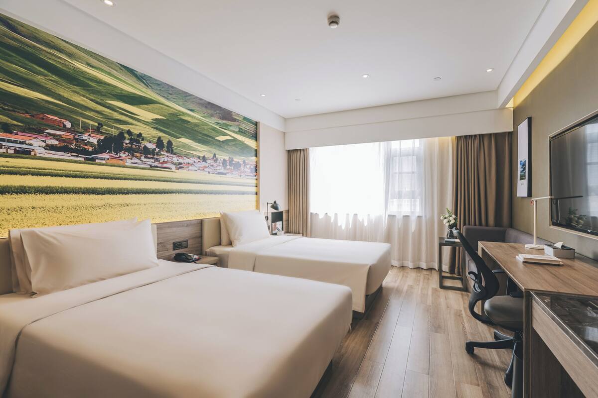 Ein Hotelzimmer mit zwei Betten, einem Schreibtisch, einem Stuhl, einem Fernseher und einem großen Fenster mit Vorhängen.