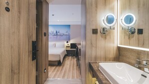 A modern hotel room with a wooden floor, a large bed, a desk with a chair, a bathroom with a large mirror and sink, and a cityscape painting on the wall. - Echarm PLUS Hotel Chongqing  (Chongqing)
