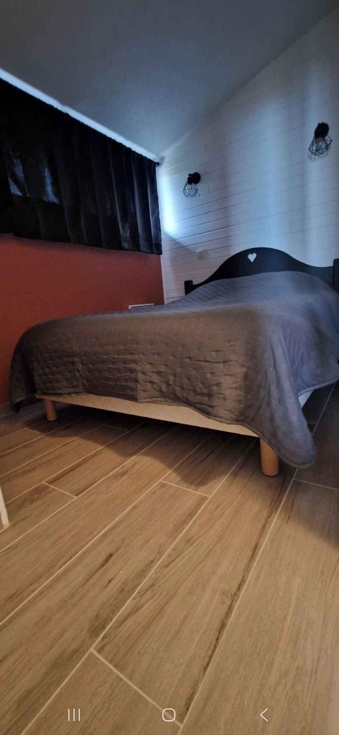 Un dormitorio con cama, piso de madera, pared roja y cortina negra.