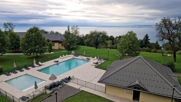 A pool area with lounge chairs and umbrellas, surrounded by a fence, with a view of a lake and distant buildings. - T2 with breathtaking view of Lake Geneva (Lugrin)