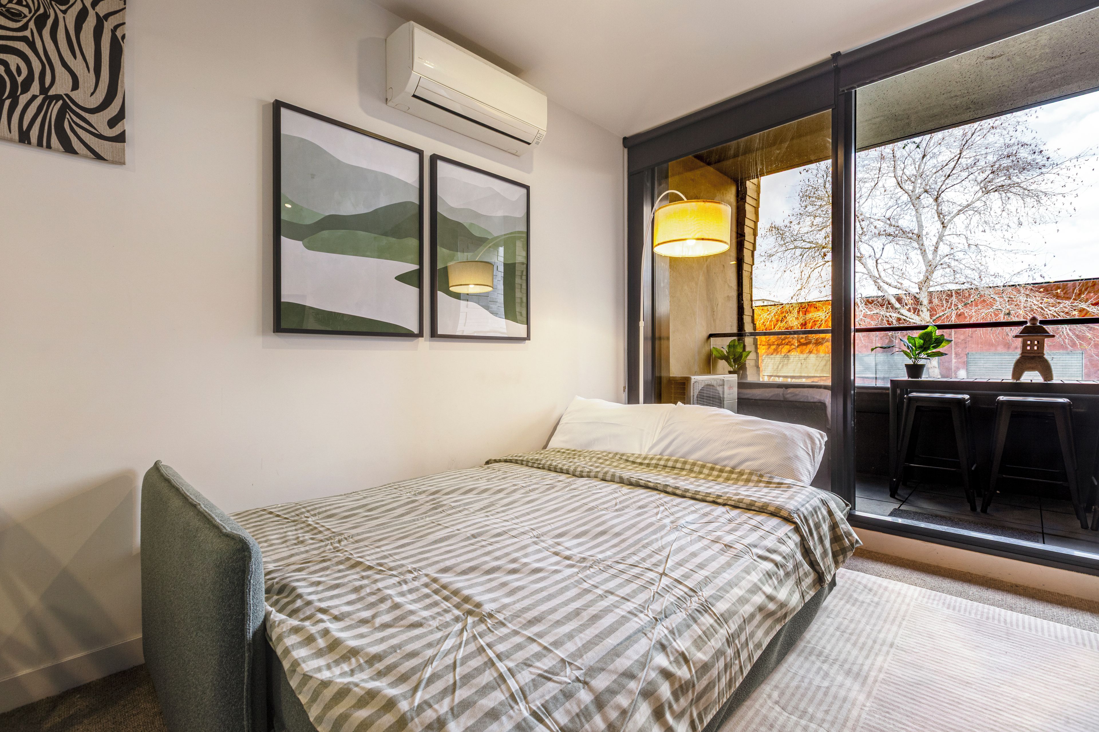 A bedroom with a bed, an air conditioner, a window with a view, and a wall-mounted lamp.