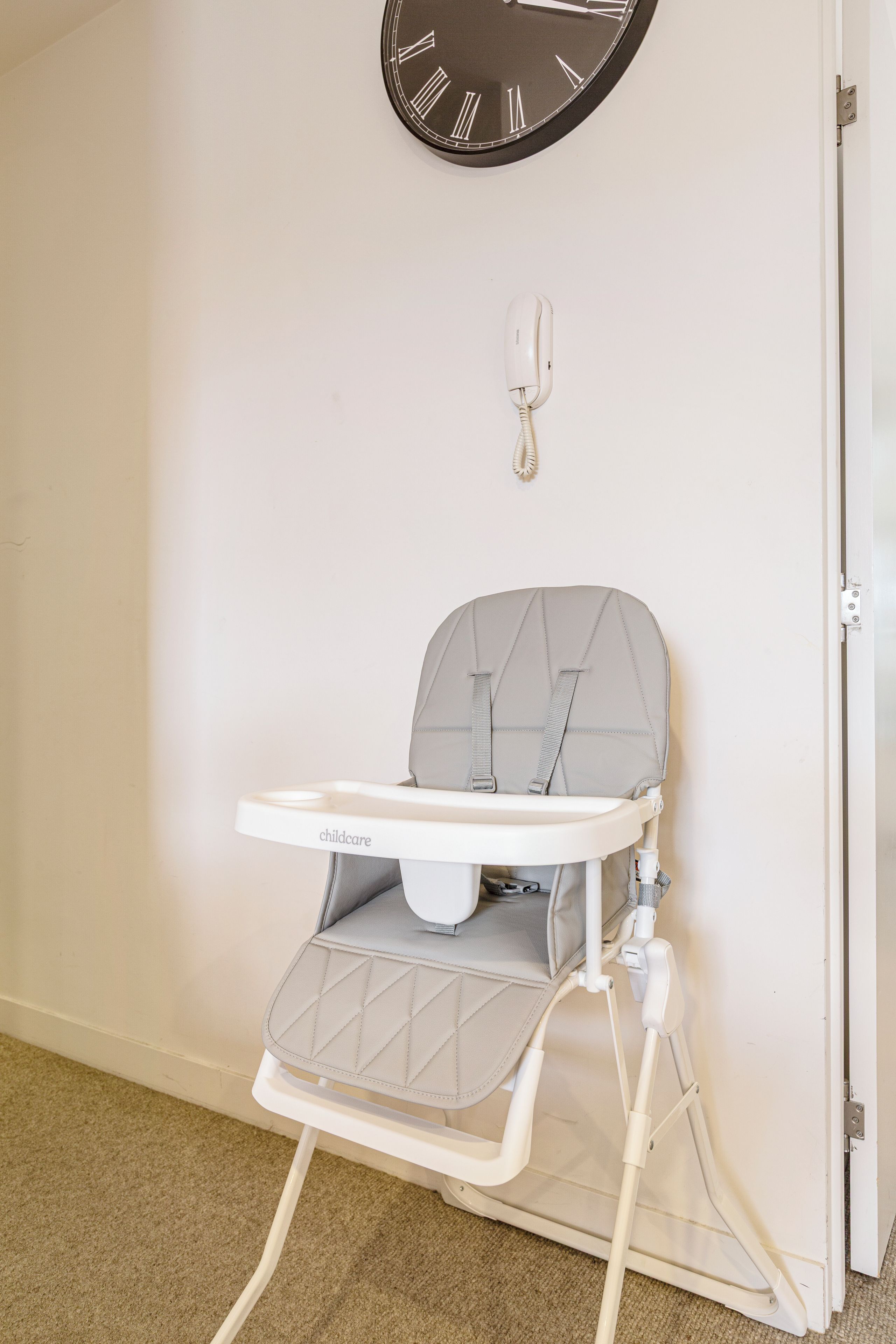 A white baby high chair with a quilted seat and backrest, positioned against a wall with a white door and a black wall clock.
