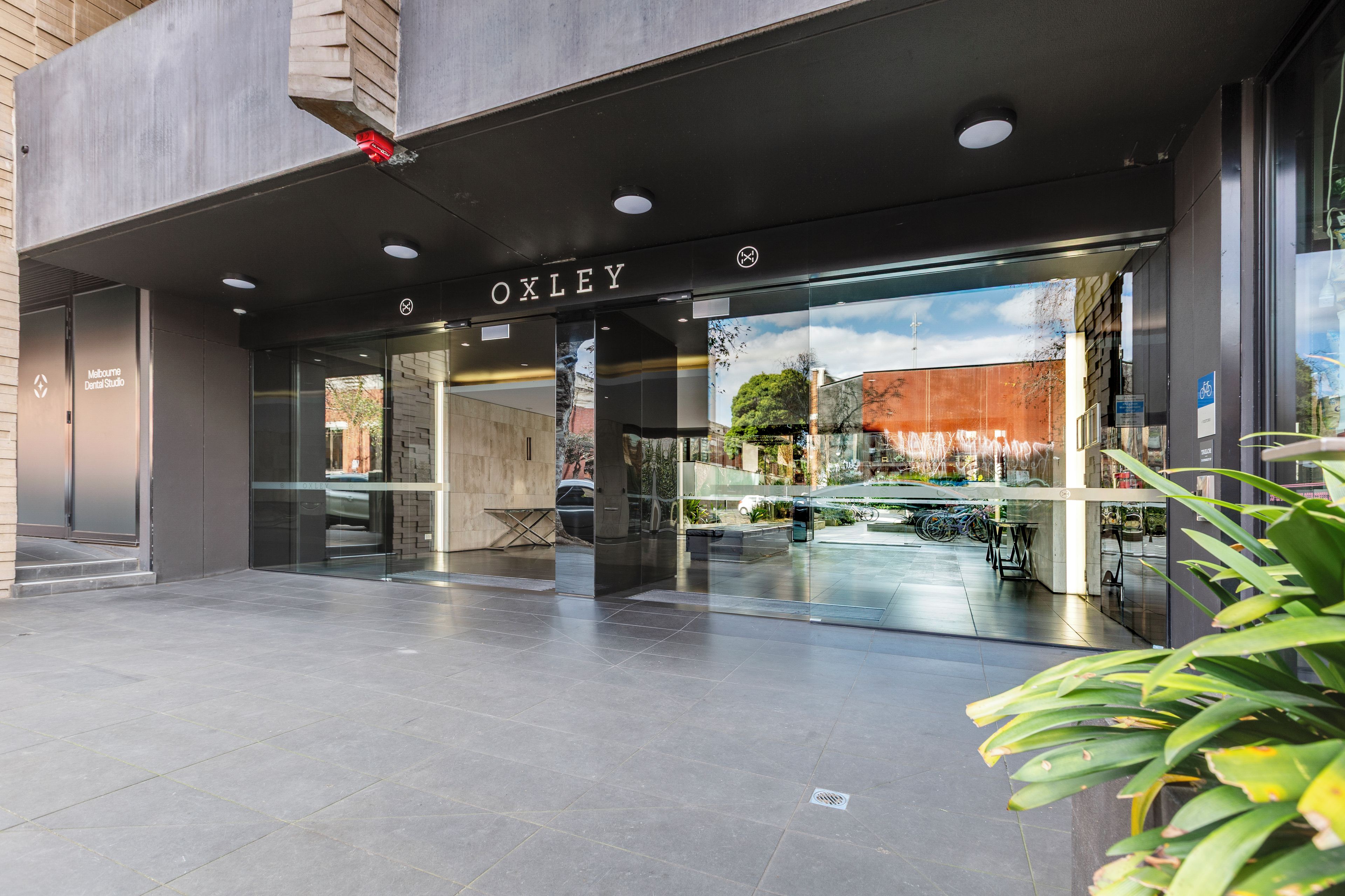 The entrance to a building with a glass facade, a sign reading 'OXLEY', and a bike parking area outside.