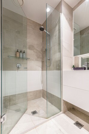 A modern bathroom with a glass shower enclosure, white tiles, and a glass shelf with toiletries. - Urban Oasis | Sleep 6 | Free Parking | Nintendo Switch (Collingwood)