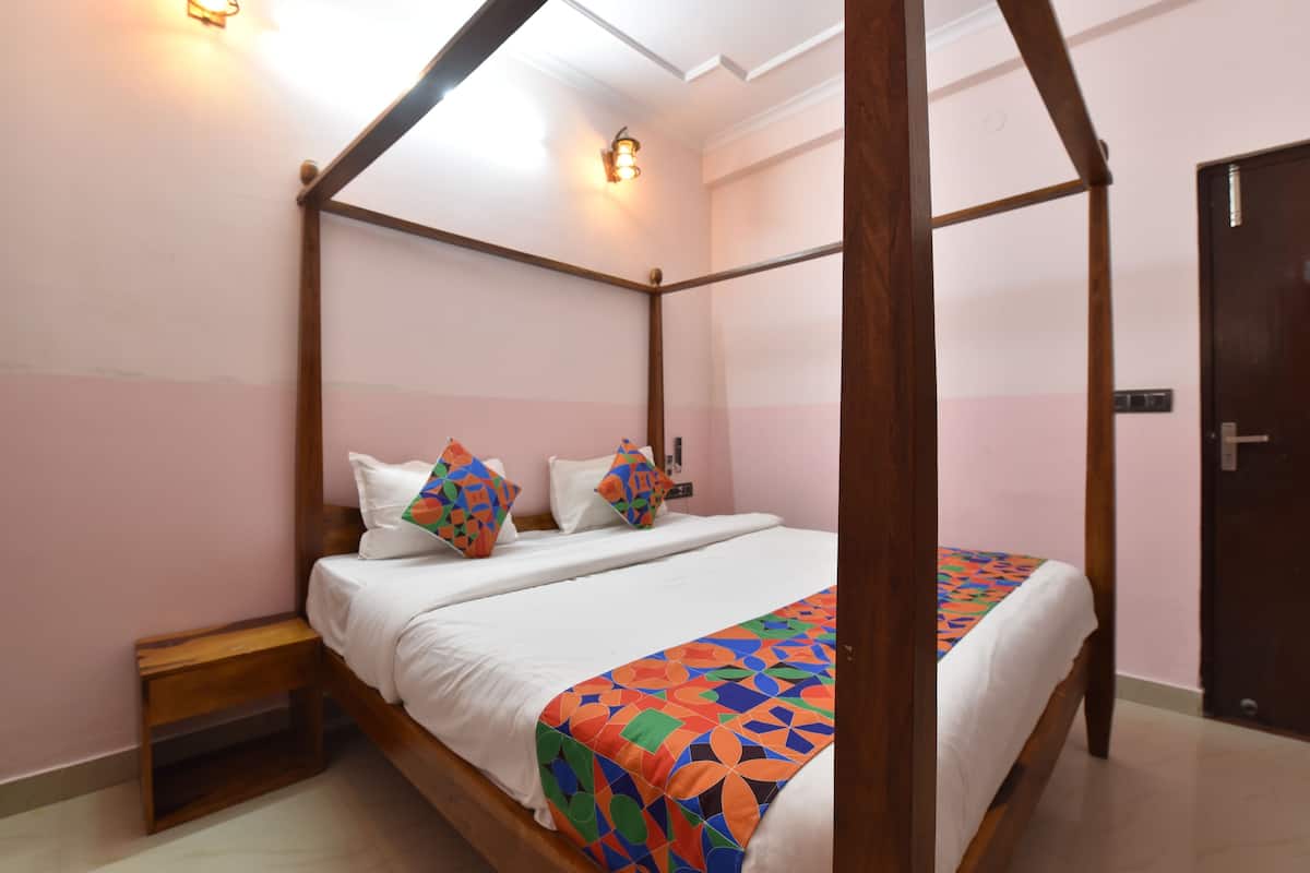 Uma cama com dossel, roupa de cama branca e almofadas coloridas, uma mesa de cabeceira de madeira e uma porta à direita.