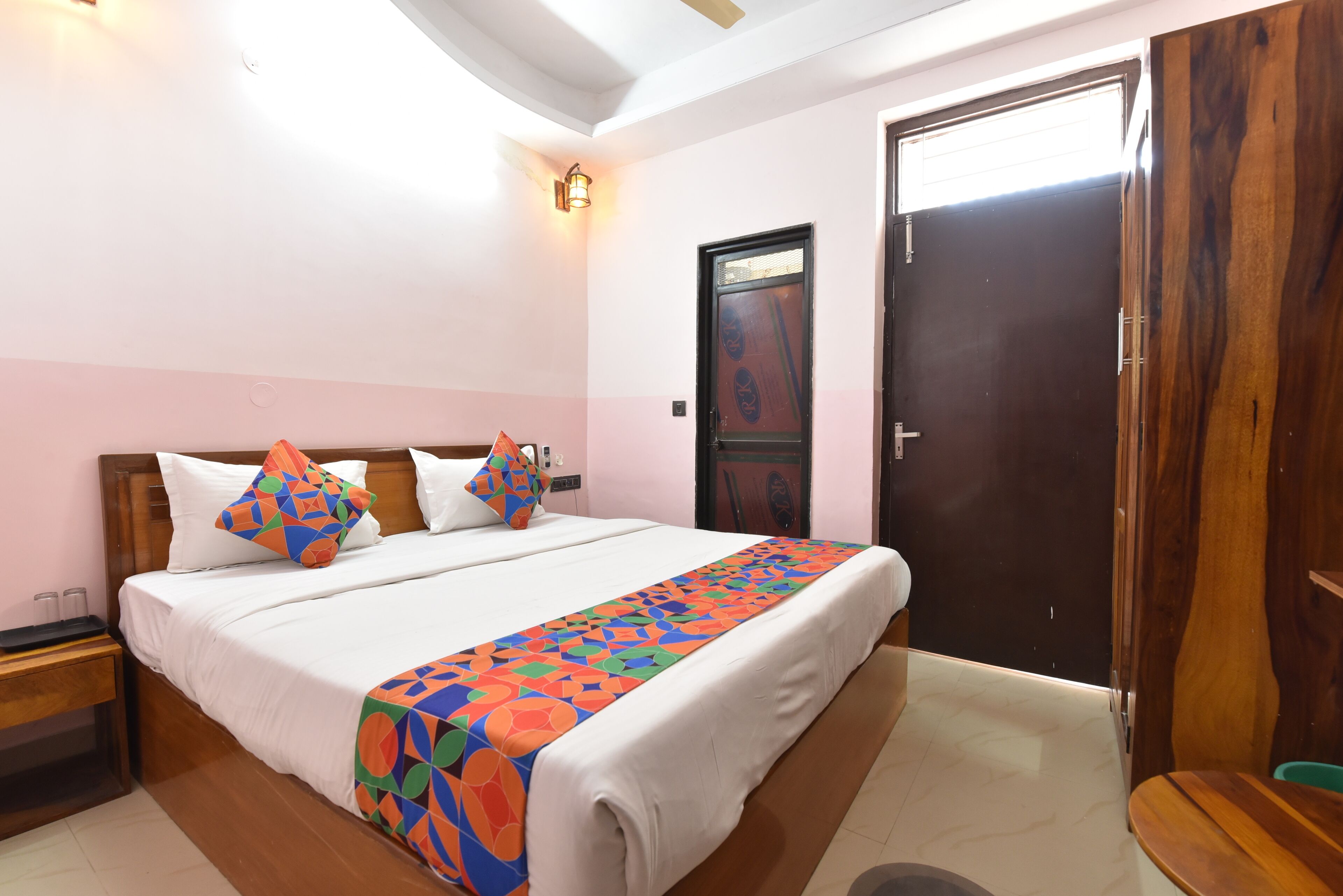 A bedroom with a bed, pillows, a nightstand, a wooden door, and a wall-mounted light fixture.