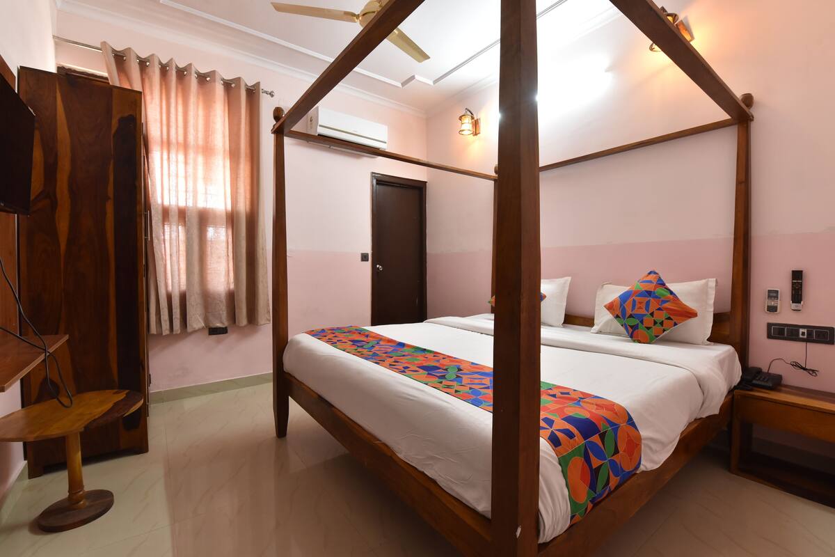 Uma cama com dossel e um edredom colorido, uma mesinha de cabeceira de madeira, uma escrivaninha com cadeira e um ventilador de teto.