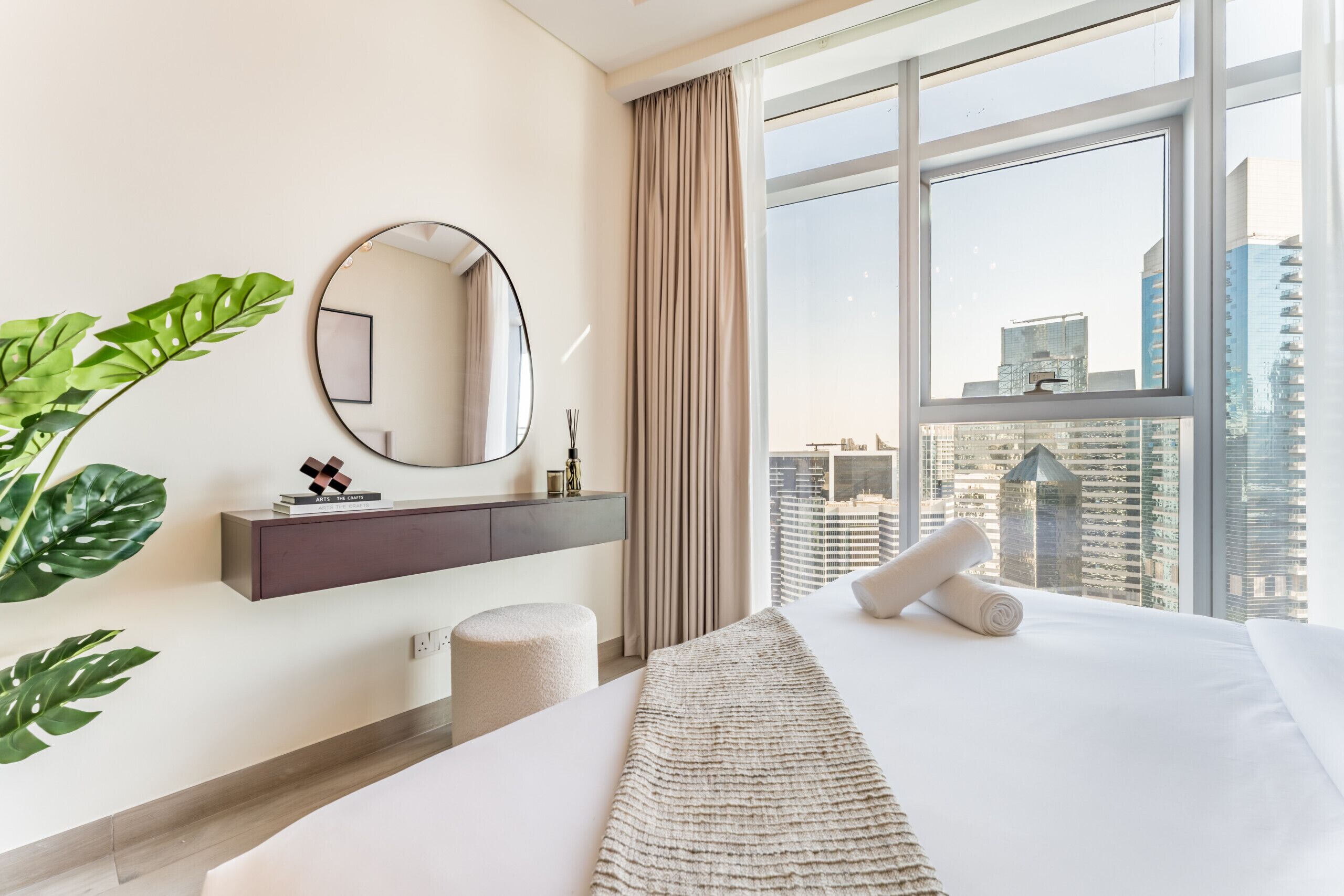 A modern hotel room with a large bed, a round mirror, a plant, and a view of tall buildings.