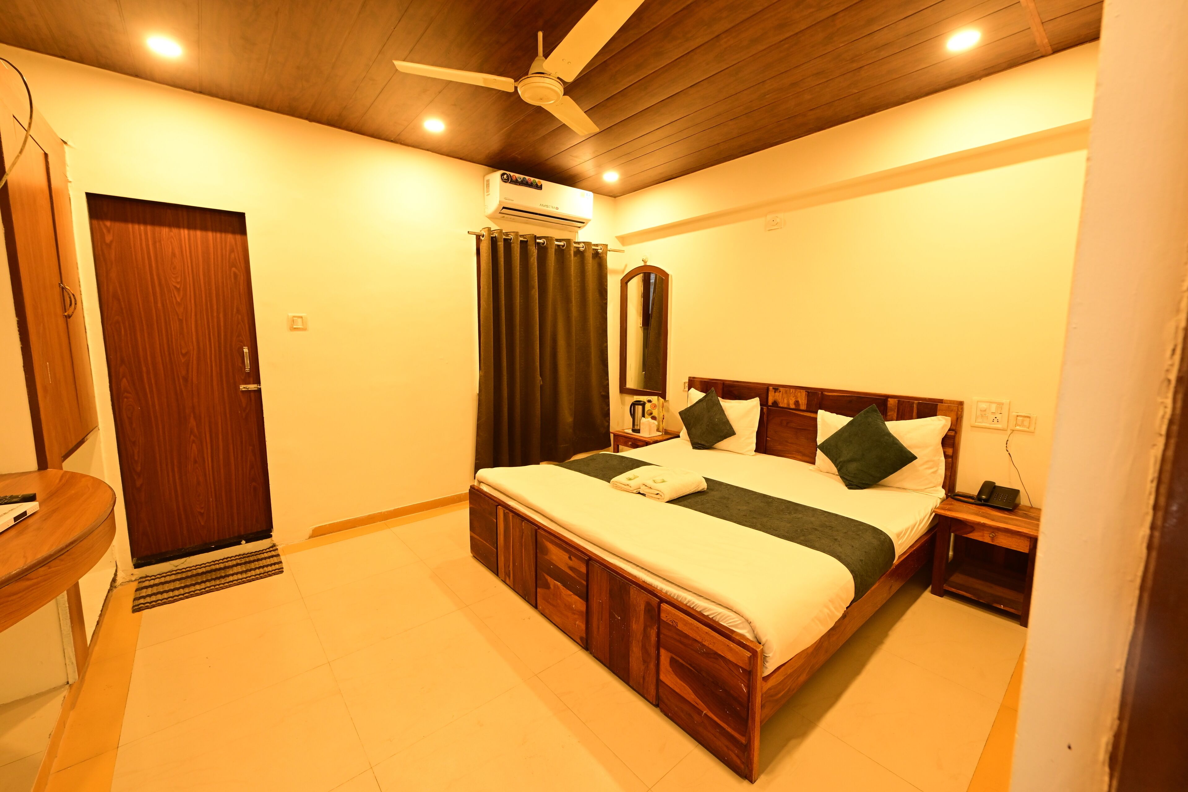A bedroom with a wooden bed, a ceiling fan, an air conditioning unit, a bedside table with a lamp, and a door.