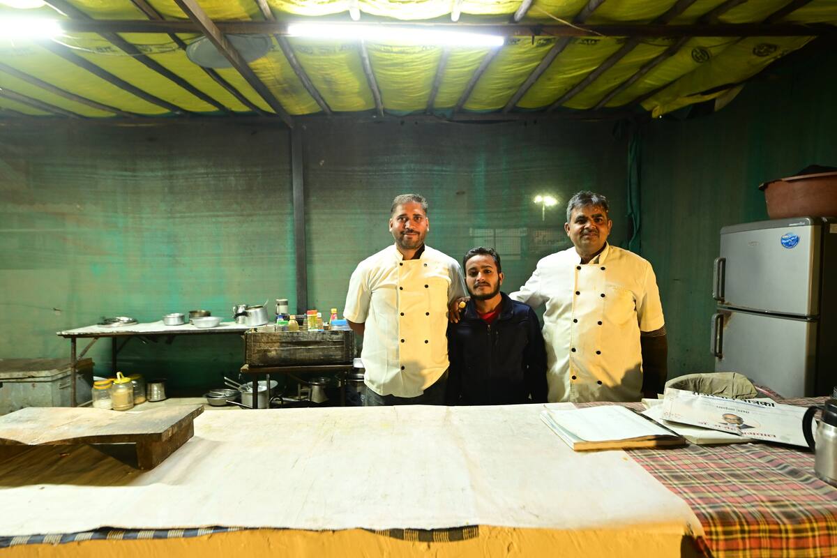 Three chefs standing in a kitchen with a table, cooking utensils, and a refrigerator.