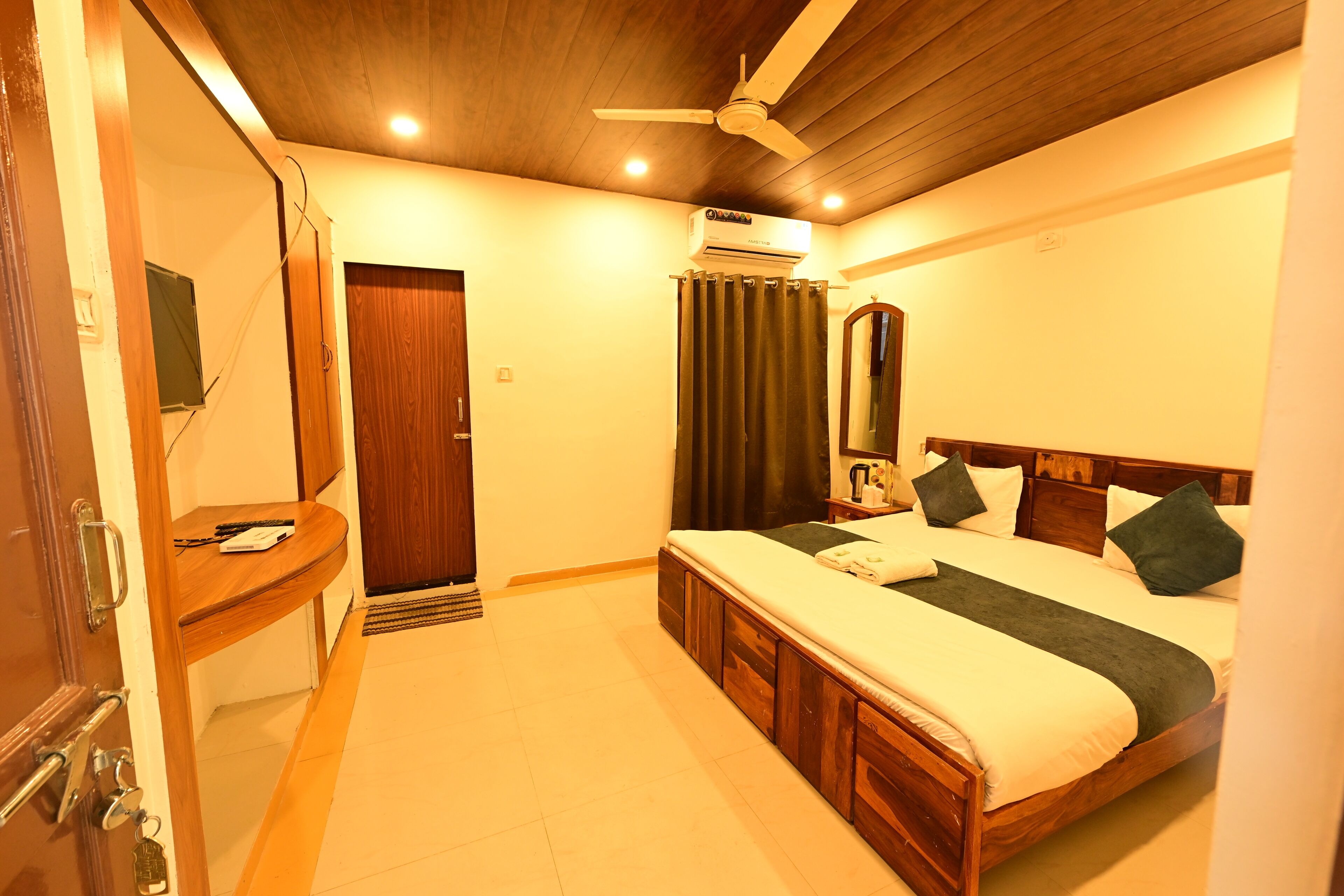 A bedroom with a wooden bed, a ceiling fan, an air conditioning unit, a wooden desk, and a door.