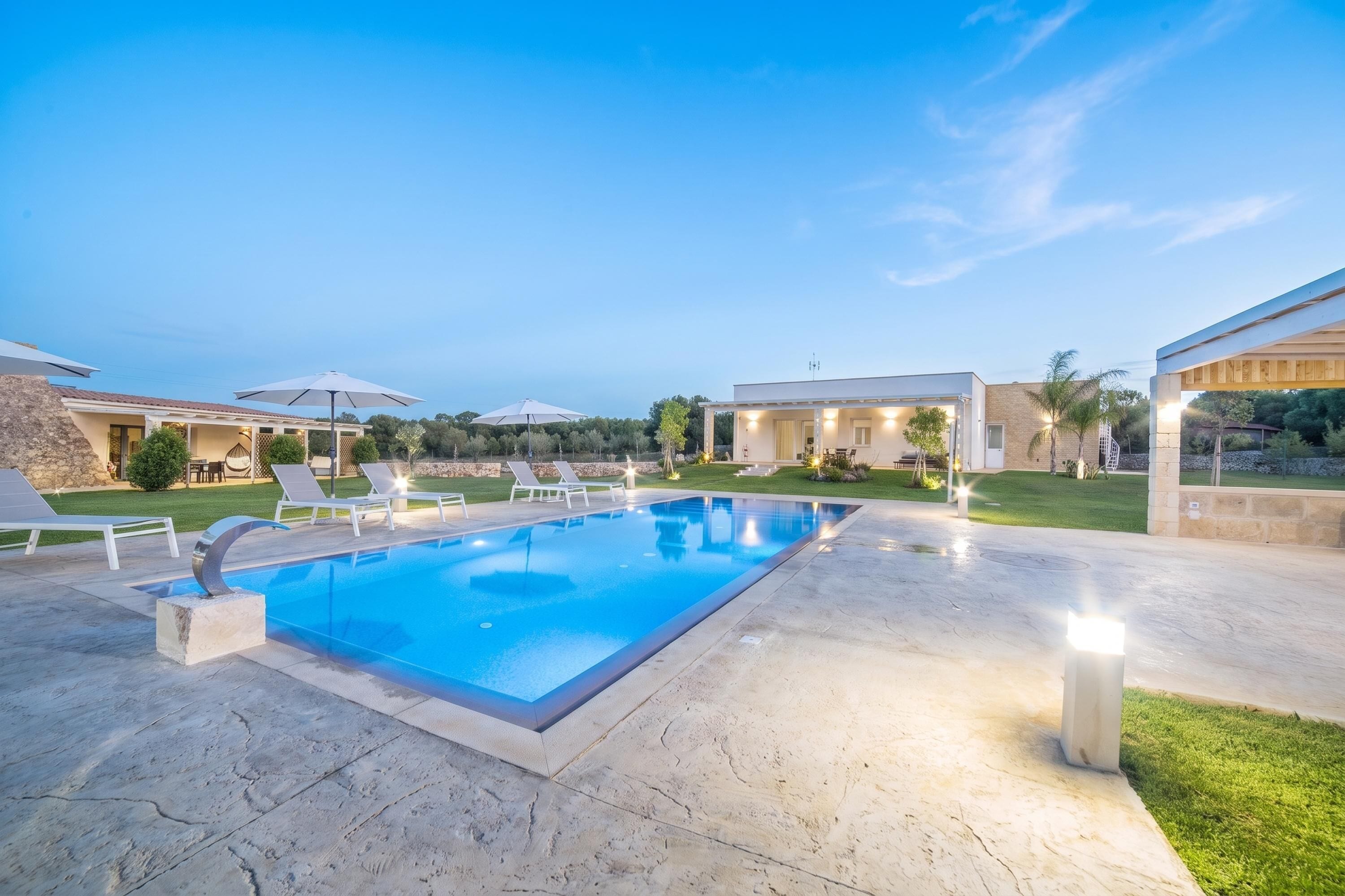 A swimming pool with lounge chairs and umbrellas, surrounded by a landscaped garden and a building with a covered patio.