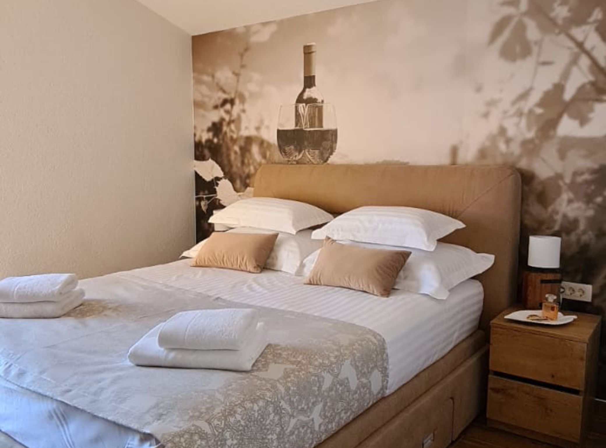 A neatly made bed with white and beige linens, a wooden headboard, and a nightstand with a lamp and a bottle.