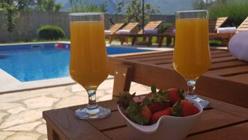 Two glasses of orange juice on a wooden table with a bowl of strawberries, near a pool with lounge chairs and mountains in the background.