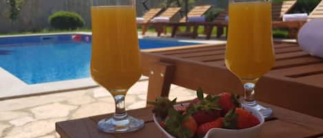 Two glasses of orange juice on a wooden table with a bowl of strawberries, near a pool with lounge chairs and mountains in the background.