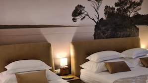A hotel room with two beds, each with white linens and brown pillows. There is a nightstand with a lamp between the beds. The wall features a large painting of trees and a rocky landscape.