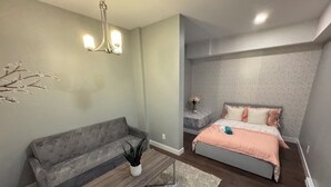 Un dormitorio moderno con un sofá gris, una cama con colcha color melocotón, una mesita auxiliar con un florero y una lámpara colgante.