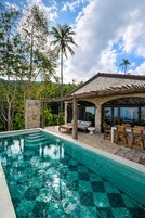 A pool area with stone walls, wooden furniture, and a view of the surrounding greenery.