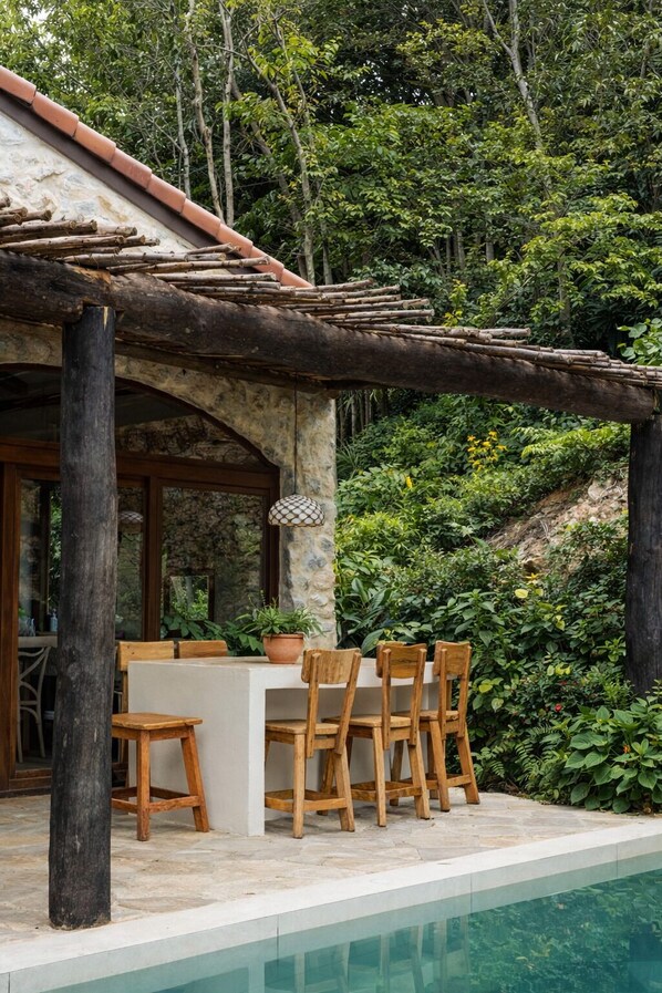 An outdoor dining area with a stone table and wooden chairs, surrounded by lush greenery and a pool. - eterna village (Ko Pha-ngan)