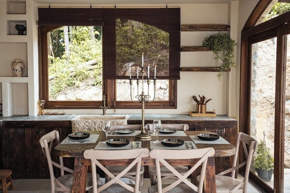 Un comedor con una mesa de madera rústica dispuesta para cuatro personas, rodeada de sillas con cojines. Un gran ventanal con contraventanas de madera oscura deja entrar luz natural y ofrece vistas al follaje exterior.