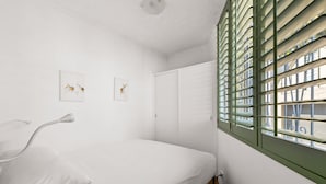 Ein schlicht eingerichteter Raum mit einem Bett, einem Fenster mit Jalousien und zwei an der Wand befestigten Kunstwerken.