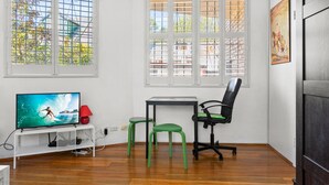 Ein Zimmer mit Fernseher auf einem weißen Stand, einem Schreibtisch, zwei grünen Hocker, einem schwarzen Bürostuhl und großen Fenstern mit Metallgitter.