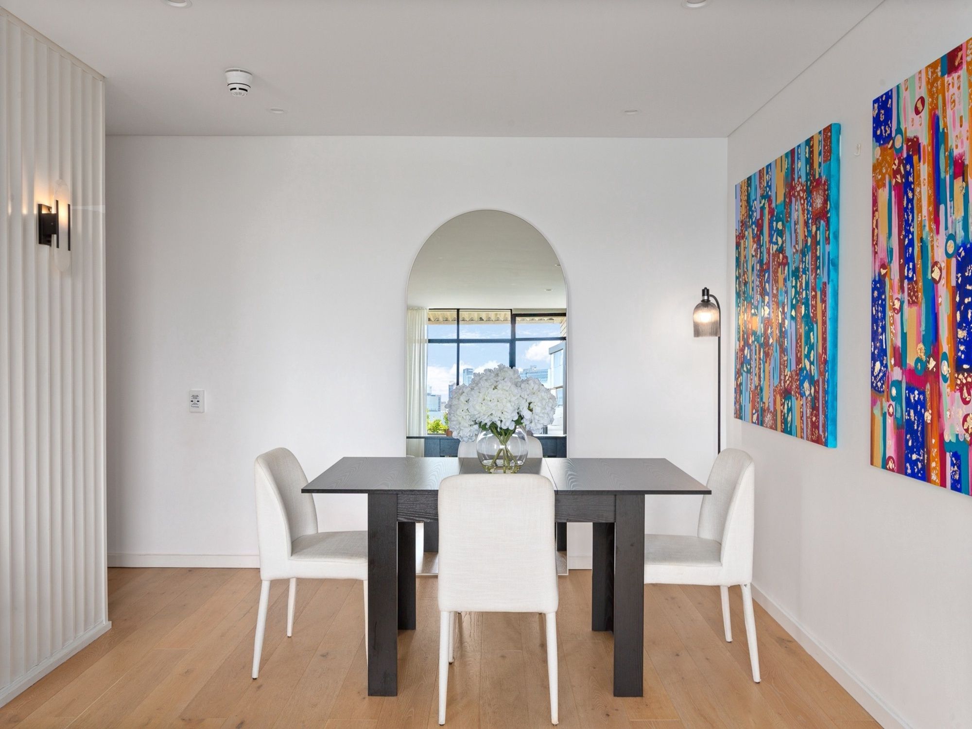 A modern dining room with a dark wooden table, white chairs, and a vase of white flowers.