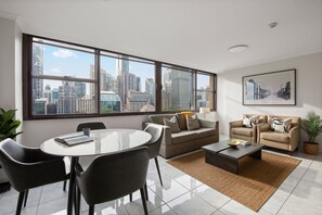 A modern living room with a dining area, large windows with a city view, a sofa set, and a coffee table. - Luxe CBD Apt | Hyde Park Views + Pool & Gym & Spa (Darlinghurst)