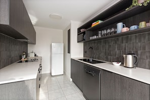A modern kitchen with dark cabinetry, a white countertop, a built-in dishwasher, and a stainless steel kettle. - Luxe CBD Apt | Hyde Park Views + Pool & Gym & Spa (Darlinghurst)