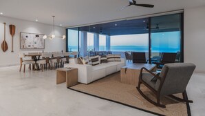 Een moderne woonkamer met een groot raam, een plafondventilator en een eethoek met een tafel en stoelen.